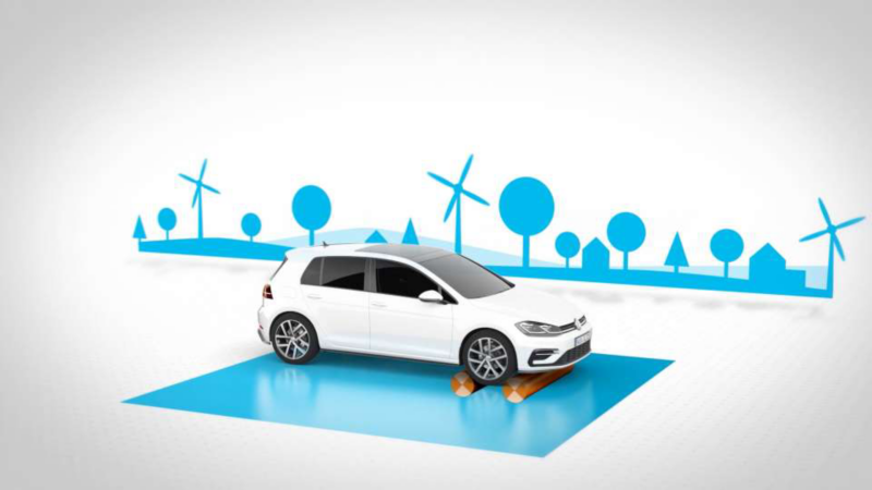 Une Volkswagen Golf blanche est présentée sur une plateforme bleue stylisée, avec un arrière-plan illustré de moulins à vent, d'arbres et de maisons, suggérant un thème écologique ou de mobilité durable.