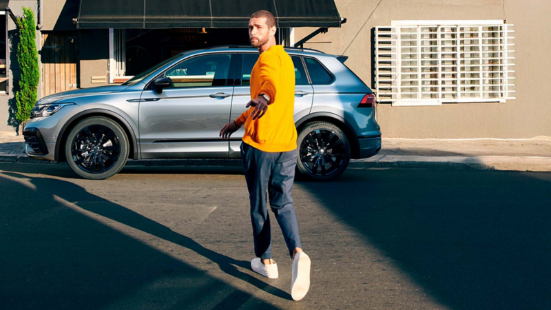 Hombre con playera amarilla camina frente a Volkswagen Tiguan 2026 color plata con rines negros estacionado en calle.