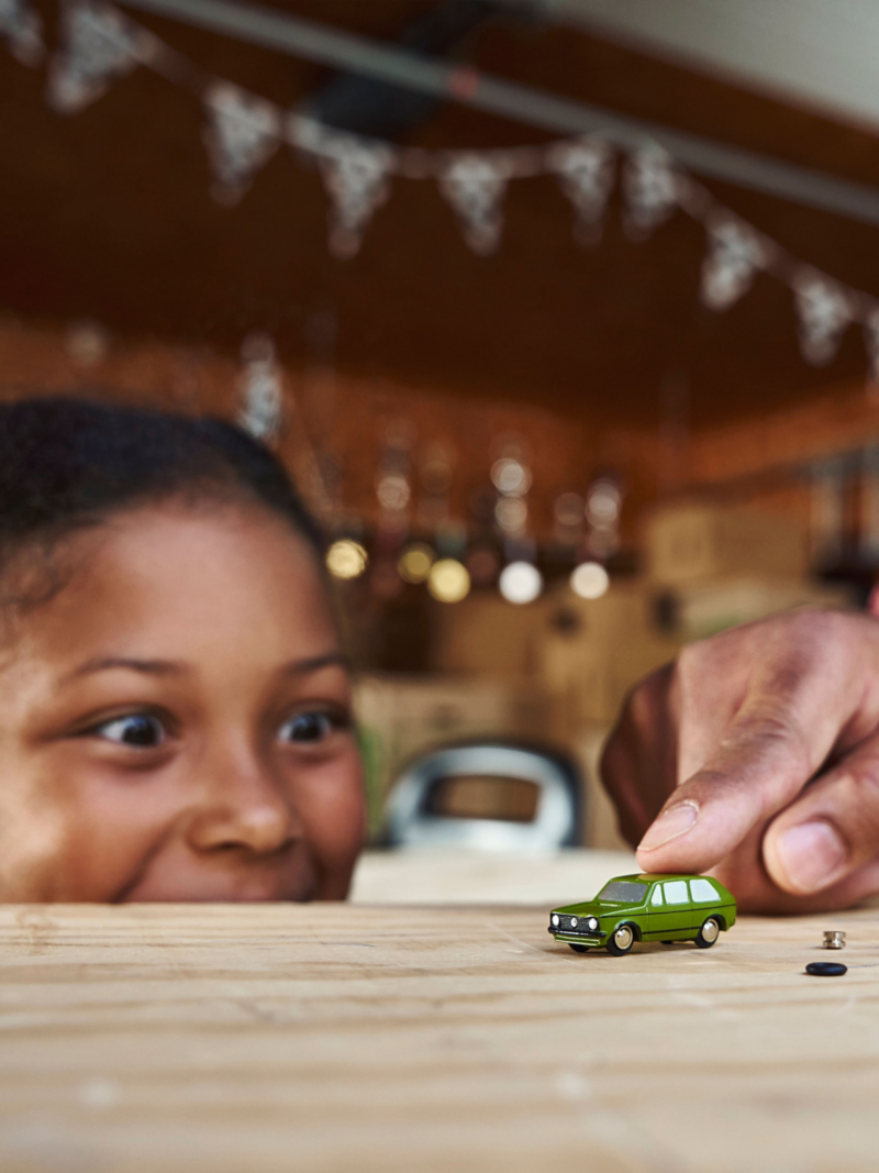 Mädchen guckt über Tischkante und beobachtet, wie jemand ein grünes Golf 1 Modellauto bewegt, daneben zwei kleine Bauteile