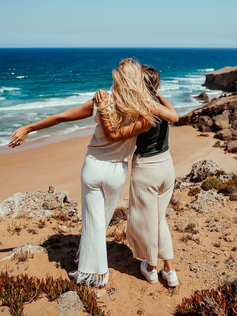 Zwei Frauen stehen Arm in Arm am Strand und blicken Richtung Meer