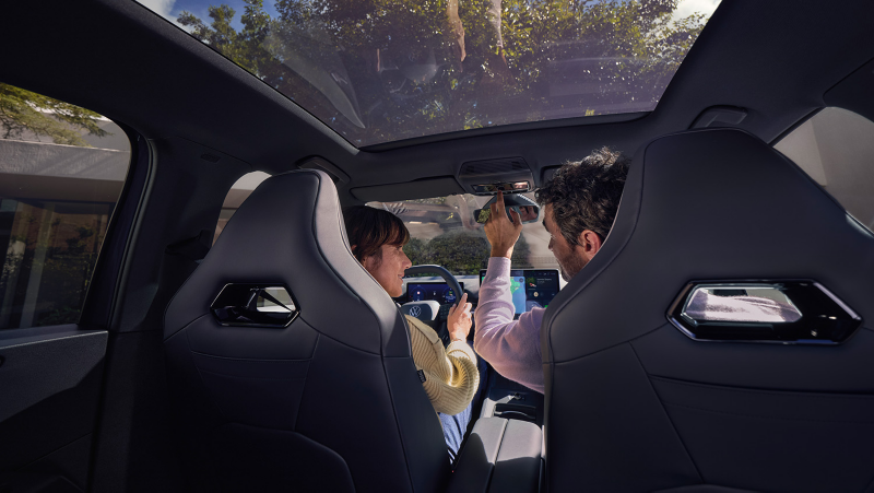 Blick von den Rücksitzen in einem VW ID.3 Neo auf zwei Personen auf den Vordersitzen, Panoramaglasdach, eine Hand greift zum Innenspiegel.