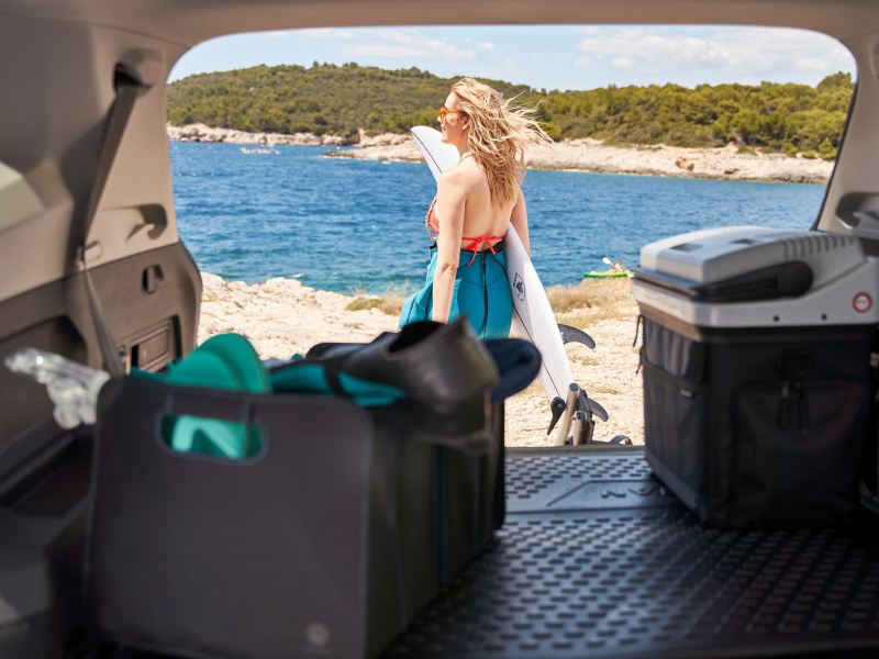 Blick aus dem Kofferraum zu einer Frau mit Surfbrett, eine Faltbox sowie Kühl- und Warmhaltebox liegen im Kofferraum