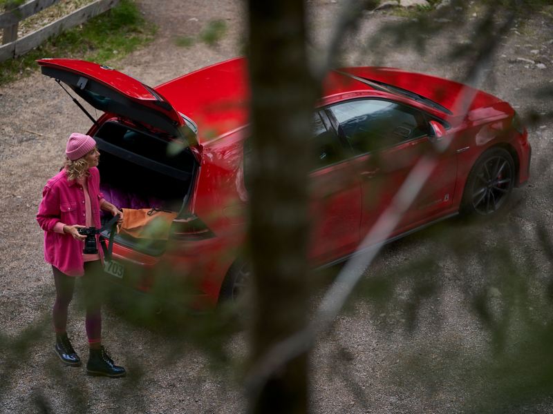 Eine Frau mit Kamera neben ihrem roten VW Golf GTI mit geöffnetem Kofferraum