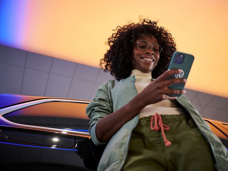Eine Frau lehnt an ihrem VW Auto und hält ein Smartphone in der Hand