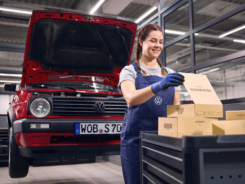 Diversos recambios Volkswagen en un taller y una persona empleada de un Servicio Oficial Volkswagen junto a un vehículo Volkswagen en primer plano