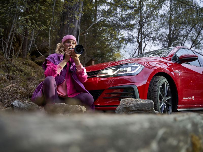 Eine Frau mit Spiegelreflexkamera hockt neben einem roten VW Golf GTI mit leuchtenden Scheinwerfern
