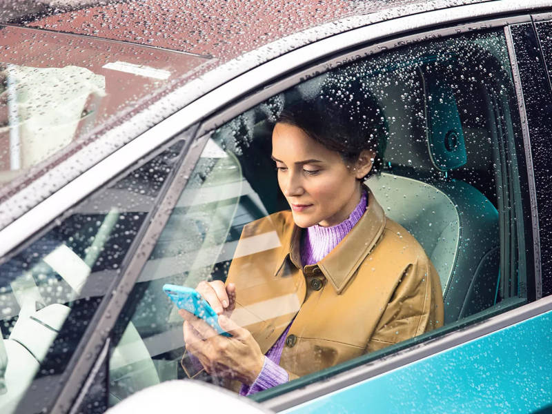  Une femme assise dans une voiture, vue à travers le pare-brise mouillé par la pluie, utilise son téléphone portable. 