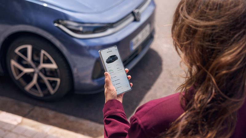 Person hält ein Smartphone mit Volkswagen‑App in der Hand, im Hintergrund die Front eines parkenden blauen VW ID.3 Neo.