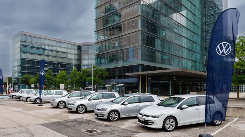 De nombreux modèles de Golf différents sont garés sur un parking devant un bâtiment VW.