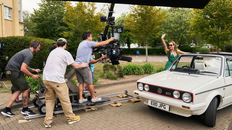 Film crew operates a camera on tracks beside a smiling woman in a white convertible. She waves energetically, evoking a joyful mood, with trees in the background.