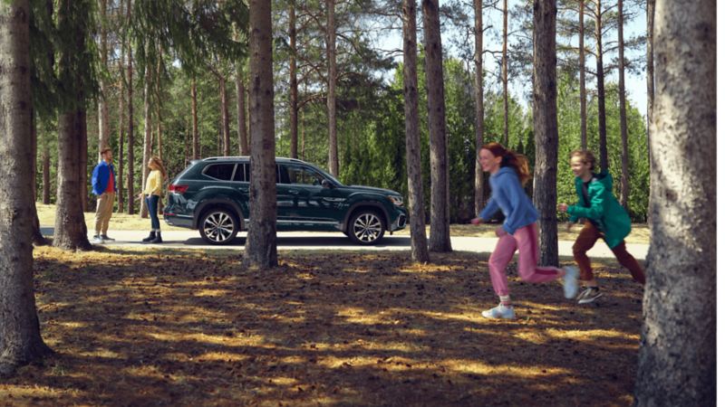 Une femme et un homme dans une forêt se tiennent debout à côté d’un VWUS Atlas 2022 de Volkswagen vert foncé métallisé pendant que leurs enfants courent autour.