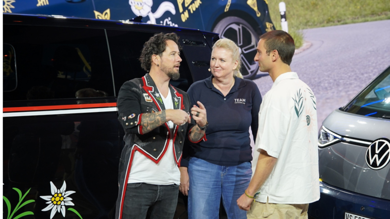 Marc Trauffer, Nicol Feissner e Pat Burgener conversano sul palco davanti a un VW Bulli dal design moderno.