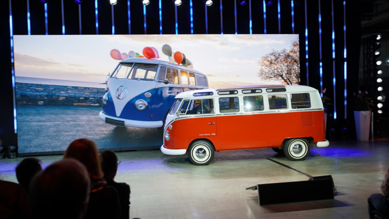 Ein roter klassischer VW Bulli steht auf einer Bühne vor einer grossen Leinwand mit einem historischen Bulli-Motiv.