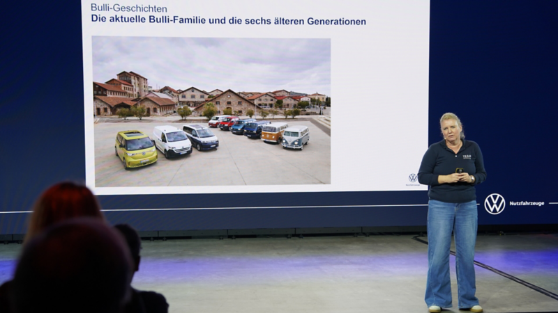 Nicol Feissner präsentiert auf einer Bühne die Geschichte des VW Bulli mit einer Übersicht der aktuellen Bulli-Familie und früherer Generationen.