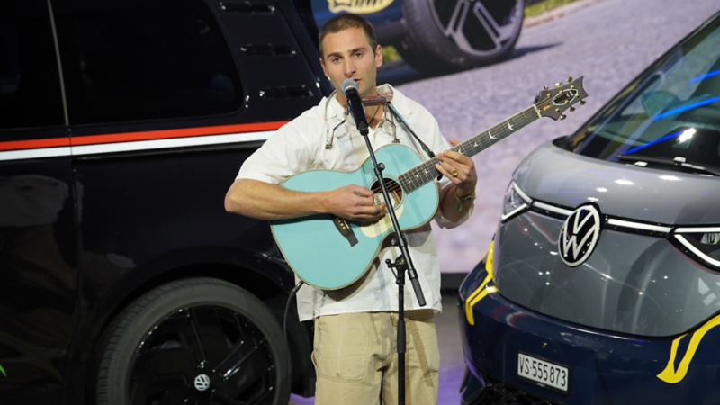 Pat Burgener suona la chitarra e canta sul palco tra due VW Bulli moderni.
