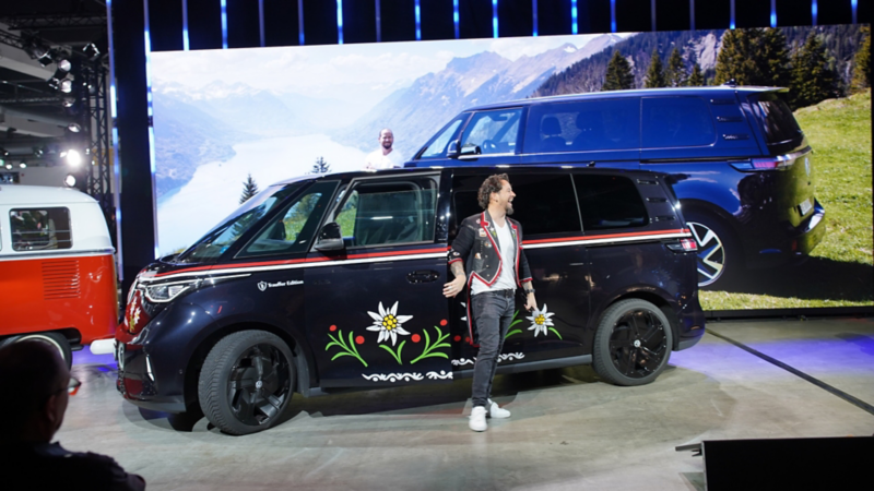 Marc Trauffer è accanto a un VW Bulli moderno personalizzato sul palco, davanti a uno sfondo alpino.