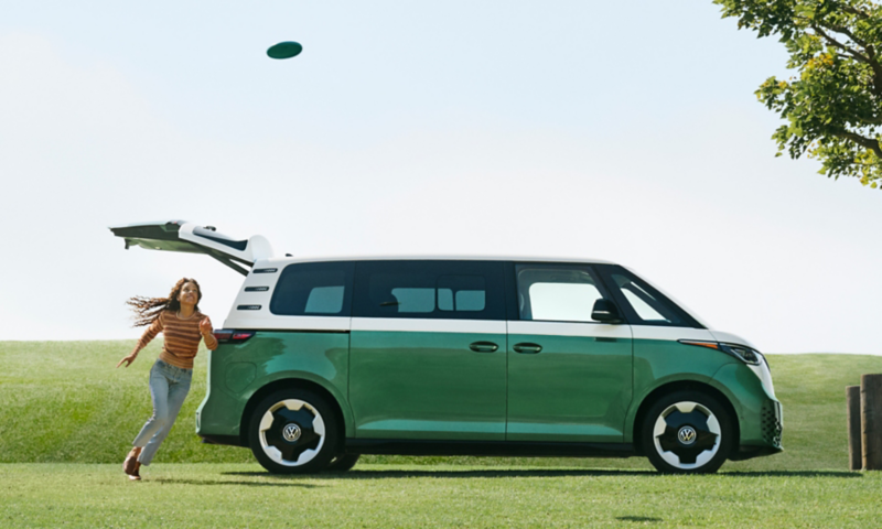 A person is playing frisbee next to an ID. Buzz EV shown in Mahi Green Metallic and Candy White near a grassy field with mountains in the background.