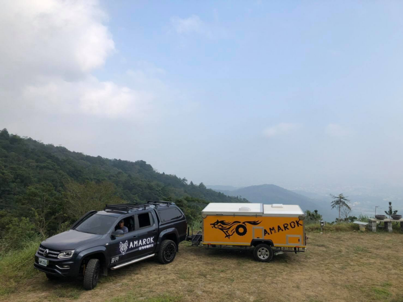 珍珠黑色Amarok拖著越野型露營拖車，停在山頂草地上，遠方低處可見城市建築物
