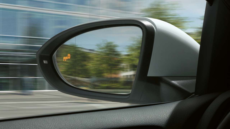 Gros plan sur le rétroviseur latéral d'une voiture, montrant un voyant d'alerte orange allumé, indiquant la présence d'un véhicule dans l'angle mort. Le reflet du paysage urbain est visible dans le miroir.