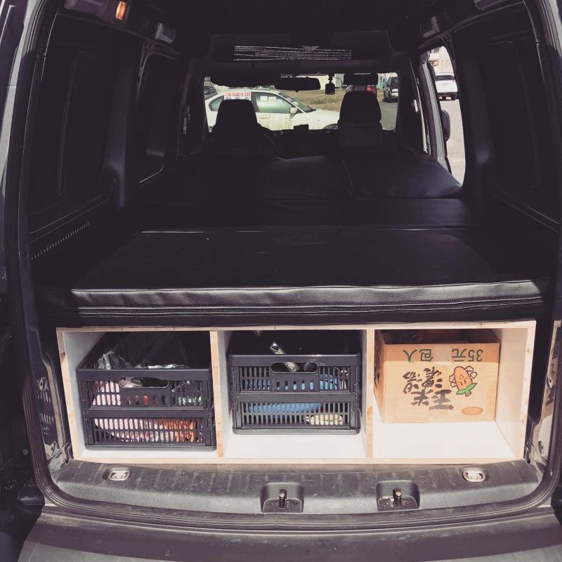 改裝的Caddy Van後廂空間，做出隔層的木板上攤平放置黑色皮墊，木板下方除了隔層、也用黑色收納籃及紙箱收納各式雜物