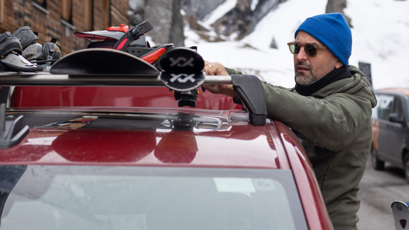 A man in a blue beanie secures skis to a red Multivan roof rack in a snowy mountain setting. He wears sunglasses and a green jacket, with other vehicles nearby.