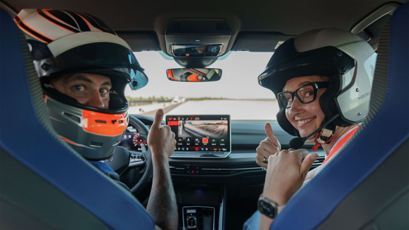Deux personnes, portant des casques de course, sont assises sur les sièges avant d'une voiture à l'intérieur bleu et noir. La personne à gauche, portant un casque gris et orange, regarde vers la caméra et fait un pouce levé. La personne à droite, portant un casque gris et des lunettes, sourit à la caméra et fait également un pouce levé. 
