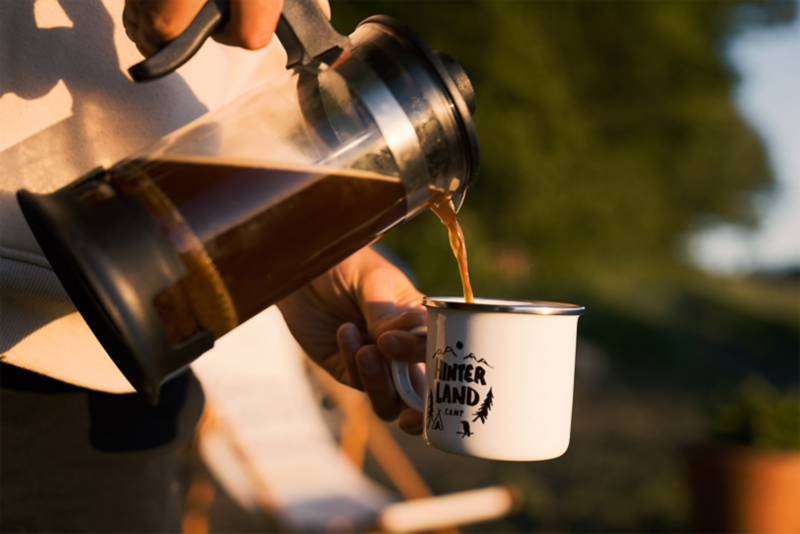 Eine Person gießt Kaffee aus einer French Press in einer mit „Hinterland“ gebrandeten Tasse ein. Die Sonnenstrahlen erzeugen eine warme Atmosphäre.
