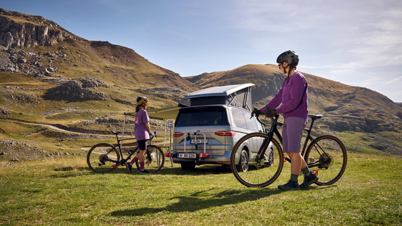 Två cyklister framför en VW California med ett soligt bergspanorama.