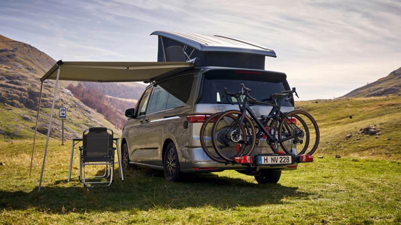 Rear view of a VW California with the awning extended and two bicycles, parked on a green mountain meadow.