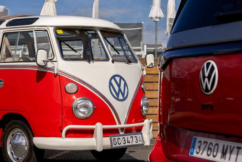 Una furgoneta clásica Volkswagen T1 'Samba Bus' de color rojo y blanco vista de frente, con su logo azul, aparcada junto a la parte trasera de una furgoneta Volkswagen moderna de color rojo, mostrando su logo y matrícula.