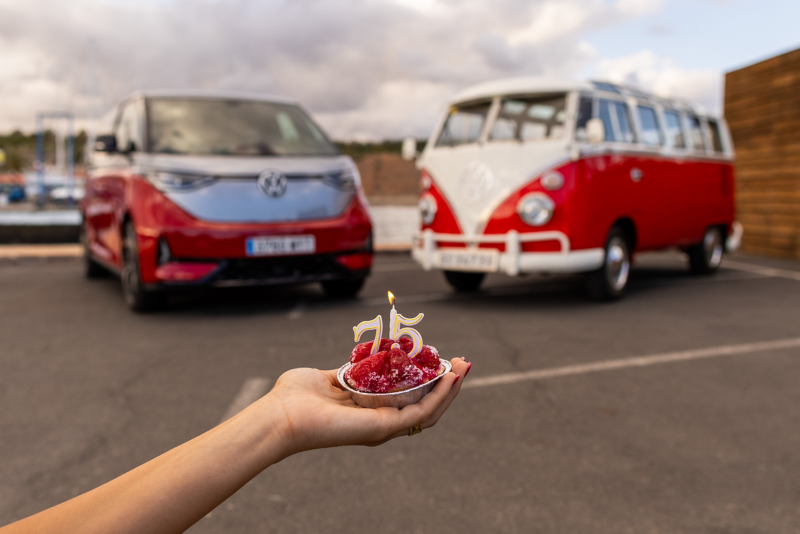 Una mano en primer plano sostiene una pequeña bandeja plateada con frambuesas rojas y velas de número 75, mientras que al fondo, desenfocadas, se ven una furgoneta Volkswagen ID. Buzz moderna roja y plateada, y una Volkswagen T1 clásica roja y blanca.