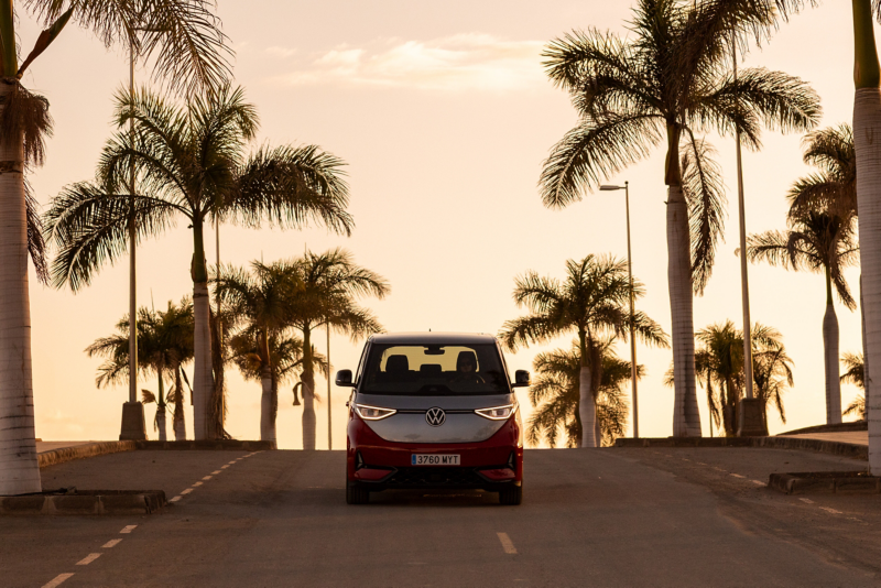 Una furgoneta Volkswagen ID. Buzz de dos tonos (rojo y plateado) vista de frente, subiendo por una carretera bordeada de palmeras al atardecer, con un cielo de tonos cálidos.