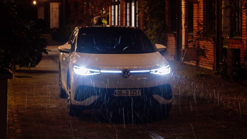 Ein weißer ID.5 steht mit eingeschalteten Frontscheinwerfern in einer Gasse, bei Nacht und Regen.