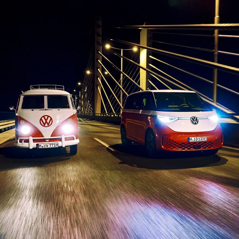 Ein VW Bulli T1 fährt neben einem VW ID. Buzz bei Nacht über eine Brücke.
