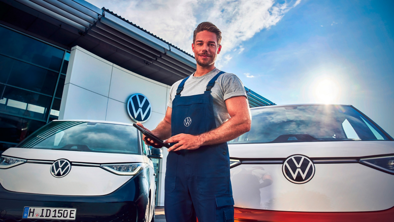 Ein VW Servicemitarbeiter mit einem Tablet in der Hand vor zwei VW Autos neben einer Werkstatt