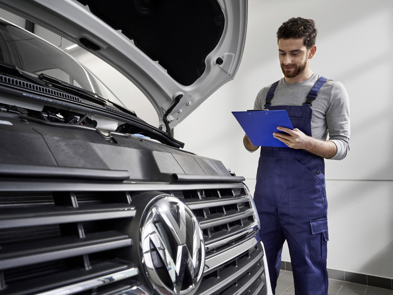 Ein Service-Mitarbeiter mit Check-Liste in der Hand überprüft den Motorraum eines VW-Fahrzeugs.