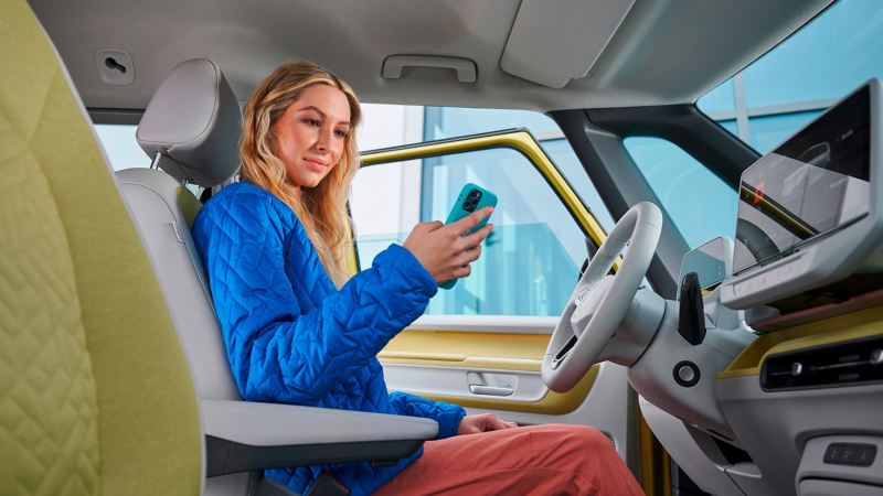 Eine Frau sitzt auf dem Fahrersitz eines ID. Buzz mit geöffneter Fahrertür und einem Handy in der Hand,