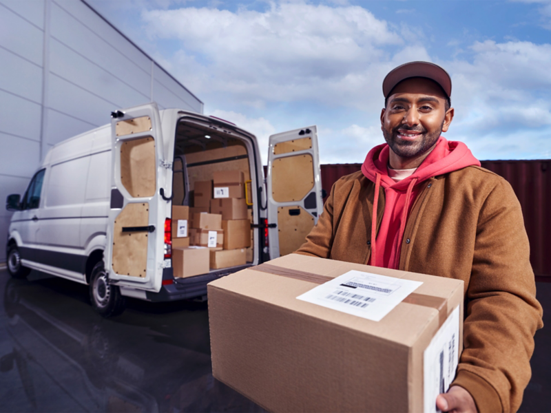 Ein Paketzusteller mit Paket in der Hand vor einem weißen VW Crafter mit geöffneter Heckklappe.