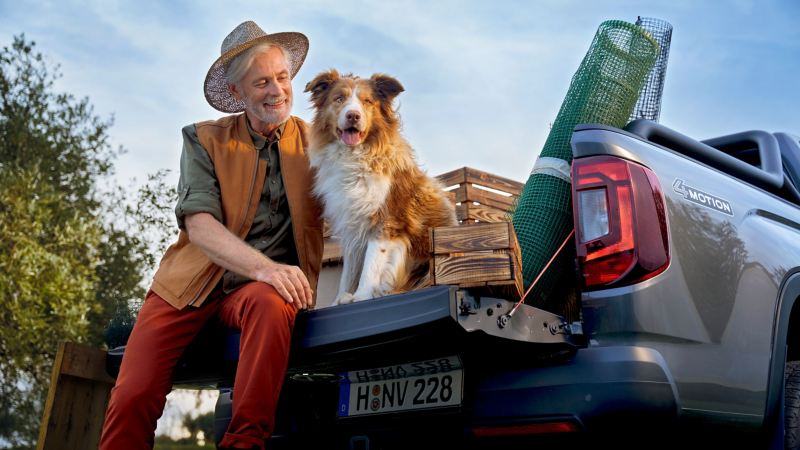 Ein Apfelbauer sitzt mit seinem Hund auf der Ladefläche eines VW Amarok. Auf der Ladefläche befinden sich Holzkisten und ein Netz für die Ernte.