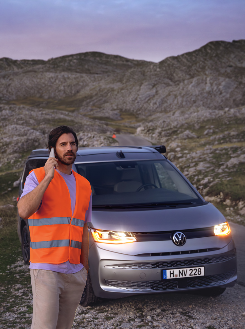 Ein Mann mit Warnweste telefoniert mit einem Smartphone, dahinter steht ein liegengebliebenes VW Nutzfahrzeug.