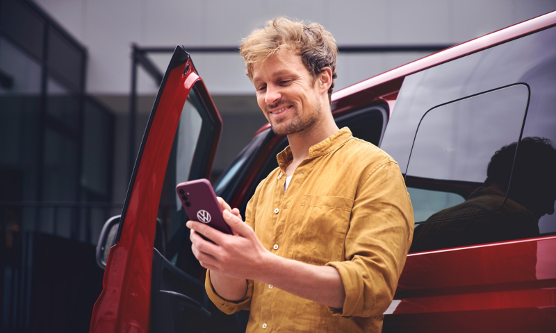 Ein Mann lehnt an einem VW Multivan und blickt auf sein Smartphone.