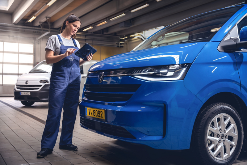 Personne en tenue de travail bleue tenant une tablette et inspectant l’avant d’un véhicule utilitaire bleu stationné dans un atelier automobile.