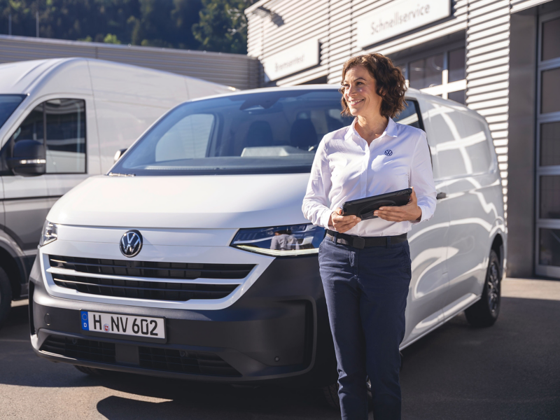 Eine Frau mit einem Tablett steht neben einem weißen VW Transporter vor der Werkstatt.