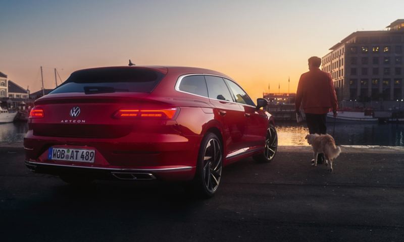 Roter VW Arteon Shooting Brake steht bei Sonnenuntergang vor einem Hafenbecken, Mann mit Hund geht darauf zu.
