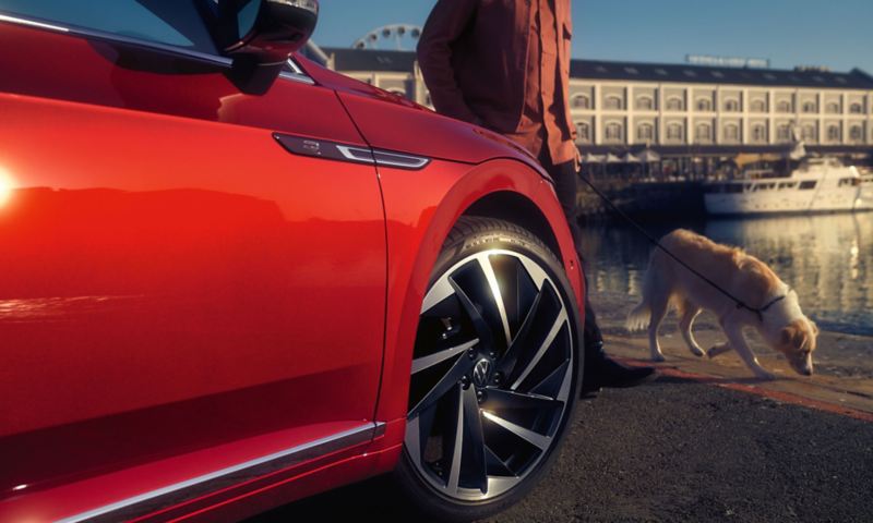 Vue détaillée de la roue avant côté passager d'une Arteon R-Line rouge avec un chien qui marche devant la voiture.