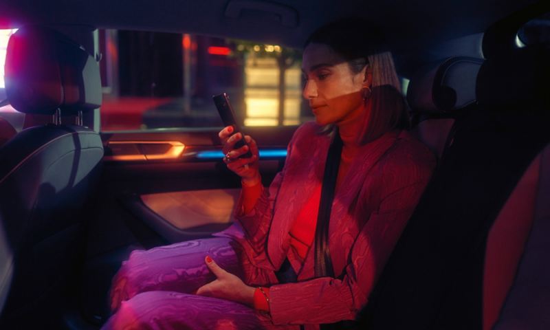 Sicht durch die offene Rücksitztür: Eine Frau im Hosenanzug sitzt auf dem Rücksitz eines VW Arteon und prüft ihr Mobiltelefon.