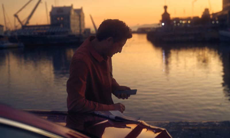 En mand læner sig op ad fronten på en rød VW Arteon og benytter sin smartphone. Aftenatmosfære på en havn.