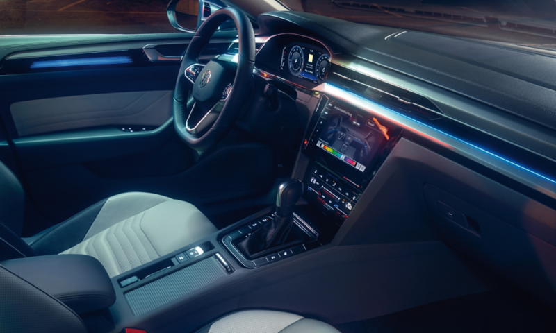 Ein Mann lehnt an der Front eines roten VW Arteon Shooting Brake und nutzt sein Smartphone. Abendliche Atmosphäre an einem Hafen.
