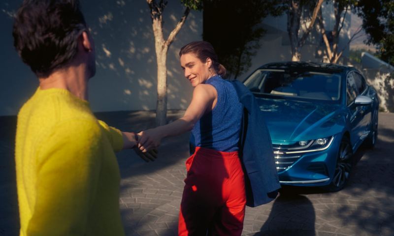 Fröhliche Frau zieht Mann am Arm zu einem blauen VW Arteon, welcher auf einer gepflasterten Auffahrt steht.