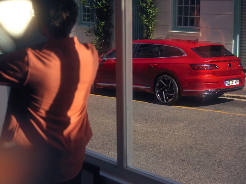 Roter VW Arteon Shooting Brake steht am Straßenrand vor einem begrünten Gebäude, Ansicht schräg hinten.